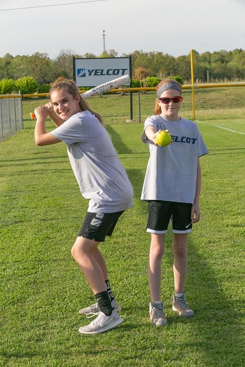 Two girls standing, one with the bat ready to swing and one holding the softball out in front of her